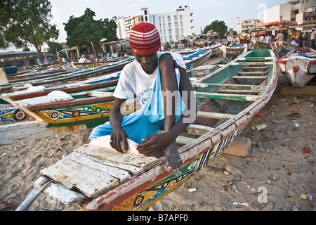 Un homme est assis sur l'un des bateaux de pêche peintes de couleurs vives qui bordent la plage à ce marché au poisson de Dakar, Sénégal. Banque D'Images