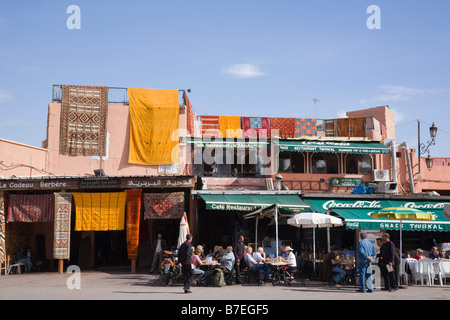 Marrakech Maroc tapis berbères étendus dehors une boutique avec les gens en ville au café de la chaussée en Place Jemma el Fna Banque D'Images