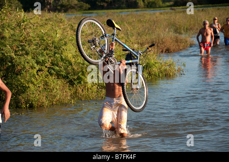 Un homme porte un vélo les gens à pied dans l'eau pour atteindre une plage de la rivière Banque D'Images