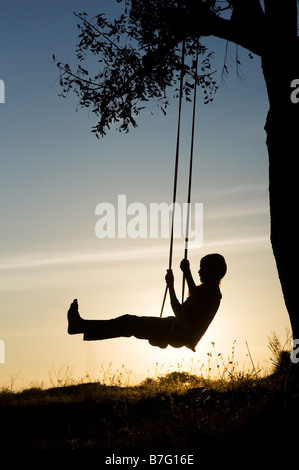 Silhouette d'Indian girl swinging sur une balançoire faite maison dans la campagne de l'Inde rurale au coucher du soleil. L'Andhra Pradesh, Inde Banque D'Images