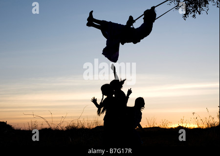 Silhouette de filles indiennes se balancer sur une balançoire faite maison dans la campagne de l'Inde rurale au coucher du soleil. L'Andhra Pradesh, Inde Banque D'Images