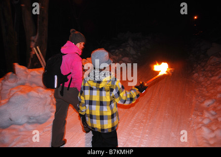 Femme adulte et jeune garçon marchant sur la piste enneigée uphil éclairé aux flambeaux Banque D'Images