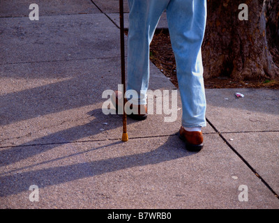 L'unstead jambes d'un homme âgé marcher sur un trottoir de la ville avec une canne, portant des jeans et des chaussures. Banque D'Images