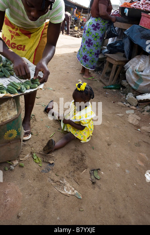 Une petite fille est assise sur le sol poussiéreux de jouer tandis que sa mère organise les marchandises sur son échoppe de marché dans la région de Poka, Nigéria Banque D'Images