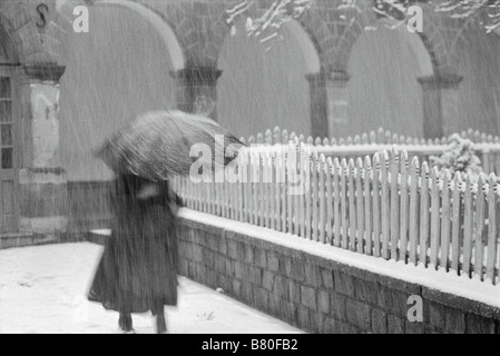 Photographie noir et blanc d'une femme marchant le long d'une clôture dans une tempête de neige humide, exerçant son activité sous un parapluie. Banque D'Images