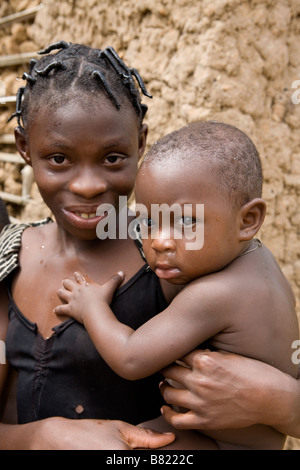 Jeune fille nigériane tient son petit frère dans le soleil à l'extérieur de leur cabane de torchis torchis de leurs village jungle à distance Banque D'Images