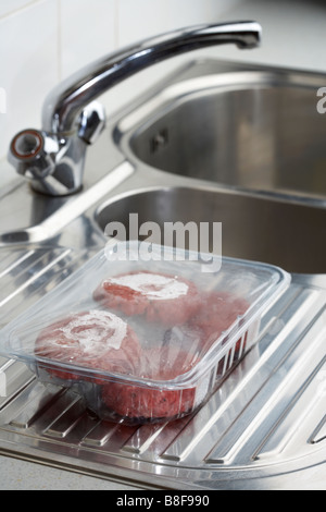 Quatre beefburgers dans un bac en plastique la décongélation à température ambiante sur une surface drainante d'évier de cuisine Banque D'Images