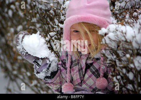 Jouent dans la neige, avec un petit sourire diabolique, une jeune fille s'apprête à lancer une boule de neige et de flocons de neige pendant l'hiver. Banque D'Images