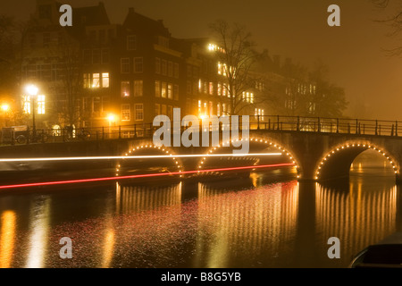 Bateau d'excursion voyages sous un pont lumineux reflète dans canal sur une nuit brumeuse à Amsterdam aux Pays-Bas Banque D'Images