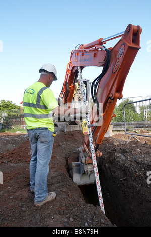 Jcb de creuser une tranchée sur un chantier tout en un ouvrier mesure la profondeur. Banque D'Images