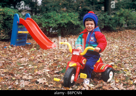 Un jeune garçon est assis sur son vélo dans sa cour pendant l'automne entouré de feuilles tombées des arbres. Banque D'Images