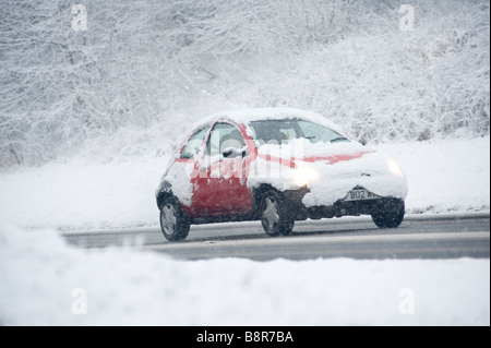 Voiture rouge conduisant lentement sur une route enneigée en hiver en Angleterre avec croisement couvertes de neige Banque D'Images