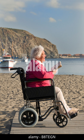 Senior satisfait personnes âgées handicapées dame assise dans son fauteuil en lisant un livre dans un endroit ensoleillé appartement de vacances plage vacances Banque D'Images