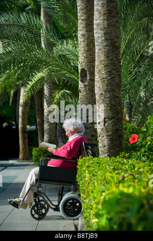 Femme âgée en vacances fauteuil roulant jardin circulation zone libre s'asseoir lisant un livre dans soleil vacances sûr vert luxuriant palmiers cadre Banque D'Images