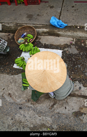 Scène de rue. Femme assise dans la rue avec Chapeau conique traditionnel de la vente de bananes dans Hoi An marché local. Vietnam Banque D'Images