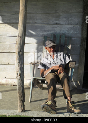 Les travailleurs plus âgés homme assis dans la chaise en bois se détendre après le travail sur le pont ensoleillé de la maison en bois simple Banque D'Images