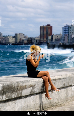 Jeune femme cubaine assis sur un mur à la mer, à la recherche dans un miroir, vérifiant son maquillage, Malecon, La Havane, Cuba, Carib Banque D'Images