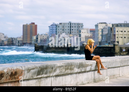 Jeune femme cubaine assis sur un mur à la mer, à la recherche dans un miroir, vérifiant son maquillage, Malecon, La Havane, Cuba, Carib Banque D'Images