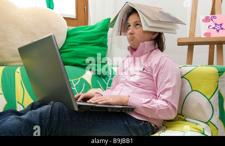 Fille de l'école, environ 11 ans, en travaillant sur un ordinateur portable et de penser avec un livre sur la tête. Banque D'Images