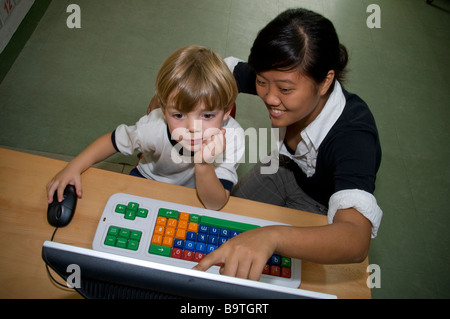 Enfant 4-6 ans en classe maternelle travaillant avec la souris sur son ordinateur à code couleur avec l'aide de son jeune professeur d'informatique à l'école Banque D'Images