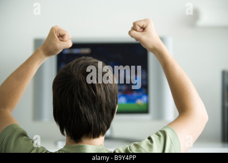 Homme regardant la télévision, poings soulevé dans l'air, vue arrière Banque D'Images