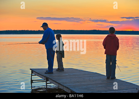 Un père avec ses deux fils de pêche à la fin d'un quai sur le lac Audy au coucher du soleil,le parc national de Riding Mountain, Manitoba,Canada. Banque D'Images