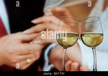 Deux verres à vin se pencher l'un vers l'autre avec Bride and Groom holding hands derrière les anneaux de mariage d'or l'affichage Banque D'Images