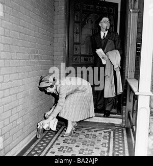 1950s, un couple quitte la maison, l'homme avec pipe et imperméable, la dame mettant des bouteilles de lait vides dans le porche couvert pour la collecte, Angleterre, Royaume-Uni. Banque D'Images