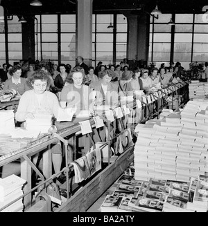 1950s, un grand groupe de femmes travaillant dans une usine d'impression assis dans des lignes, mettant des encarts ou des dépliants dans des magazines à la main, Angleterre, Royaume-Uni. Banque D'Images