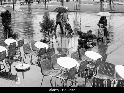 Années 1980, photo en noir et blanc, les gens d'entreprendre une marche sous la pluie, les parapluies, café-terrasse, chaises vides et plats Banque D'Images