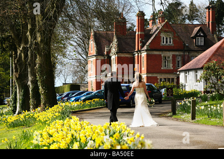 Mariée et le Marié à pied main dans la main sur le jour du mariage au printemps dans dafodills raison de campagne Salle de mariage Manor House Banque D'Images