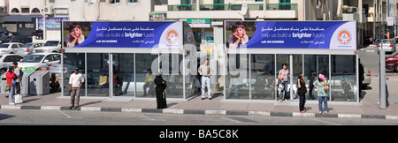 Scène de rue ensoleillée de Dubaï hommes et femmes debout à l'extérieur d'un abri de bus climatisé moderne avec panneau de publicité bilingue Émirats arabes Unis eau Banque D'Images