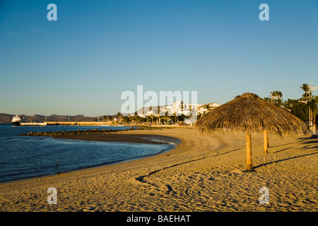 Mexique La Paz parasols en chaume Palapa Beach sur la baie le long des bâtiments de la ville et les montagnes dans la distance Banque D'Images