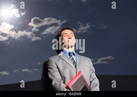 Smiling Businessman leaning against wall réfléchissante à l'extérieur, les yeux clos, holding notebook Banque D'Images