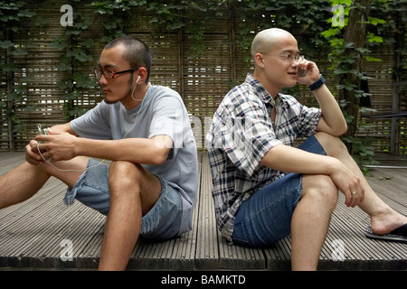 Deux jeunes hommes assis, l'un de l'écoute d'un lecteur MP3 et d'une conversation sur son téléphone cellulaire Banque D'Images