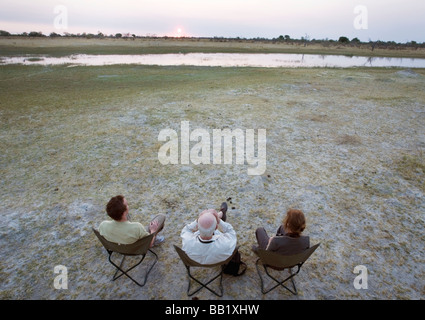 Vue grand angle de trois personnes sur safari regarder le coucher de soleil, Okavango Delta, Botswana Banque D'Images