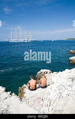 Voyages Croatie, un navire à voile mât cinq dans la mer Adriatique passe deux hommes en train de bronzer sur la côte rocheuse près de Rovinj. Banque D'Images