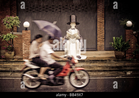 Un couple riding a motorcycle sous la pluie holding an umbrella passer une statue d'Abraham Lincoln sur le trottoir de la plage de Kata Noi Banque D'Images