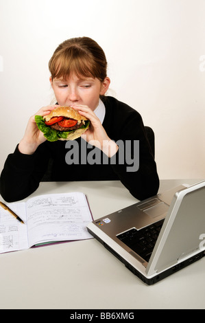 Manger une écolière beefburger avec une salade saine remplissant tout en faisant ses devoirs sur un ordinateur portable Banque D'Images