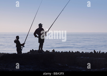 Deux pêcheurs de silhouette à l'aube la pêche au large des rochers de bord de mer. Rétro-éclairée. Silhouette. Durban, KwaZulu Natal, Afrique du Sud. Banque D'Images