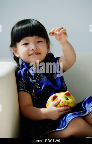 Une petite fille en bleu cheongsam soie joue avec une tirelire Banque D'Images
