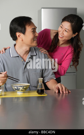 Homme mûr dans la cuisine, manger un bol de nouilles, en se tournant vers femme debout derrière lui Banque D'Images