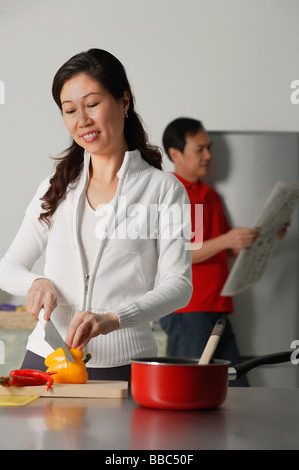 Femme mature dans la cuisine préparer un repas, l'homme en arrière-plan holding newspaper Banque D'Images