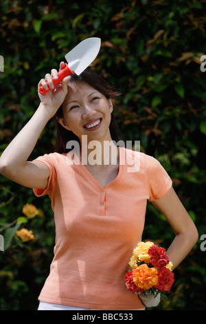 Femme tenant une truelle et bouquet de fleurs, essuyant son front Banque D'Images