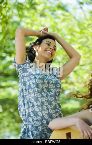 Jeune femme debout à l'extérieur avec les mains sur la tête, souriant, d'un ami qui joue de la guitare à proximité Banque D'Images