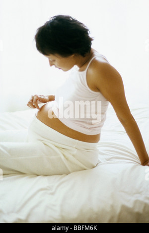 Pregnant woman sitting on bed, touchant l'estomac, side view Banque D'Images