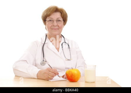 Senior female doctor sitting behind desk avec verre de lait et apple isolé sur fond blanc Banque D'Images