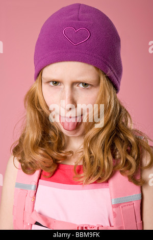 Fille aux cheveux roux portant un bonnet violet et une besace sac en face d'une toile rose, poussant sa langue maternelle
