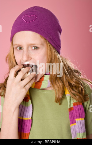 Fille aux cheveux roux portant un bonnet violet en face d'une toile rose, mordre dans une guimauve au chocolat Banque D'Images