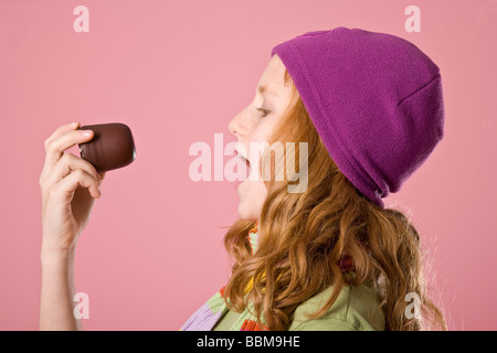 Fille aux cheveux roux portant un bonnet violet en face d'une toile rose, mordre dans une guimauve au chocolat Banque D'Images